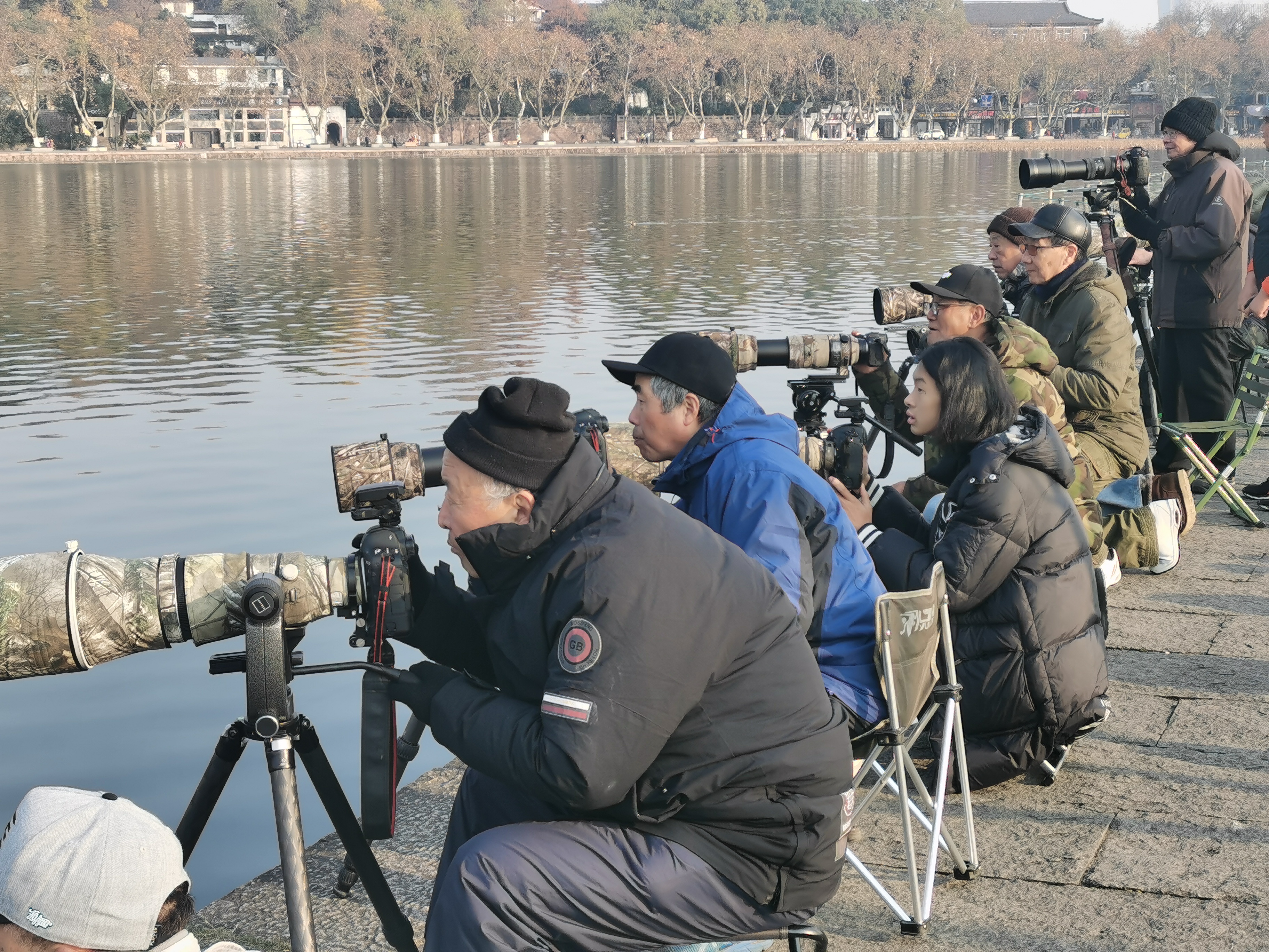 「要拍出鸕茲抓魚的大片，就要能守，抓的魚種特殊的話，盡量多按快門，往往是幾百張照片只能挑出1-2張好的。鸕茲在走之前，在西湖大量補充了自己的食慾，到了來年三月，與現在鸕茲會長的不一樣，腦袋毛色都變成了白色，所以看照片，就能看出是哪個月拍的鸕茲。」毛建平說。圖為毛建平和鳥友們等待鸕茲抓魚的一刻。（香港文匯網記者茅建興 攝）