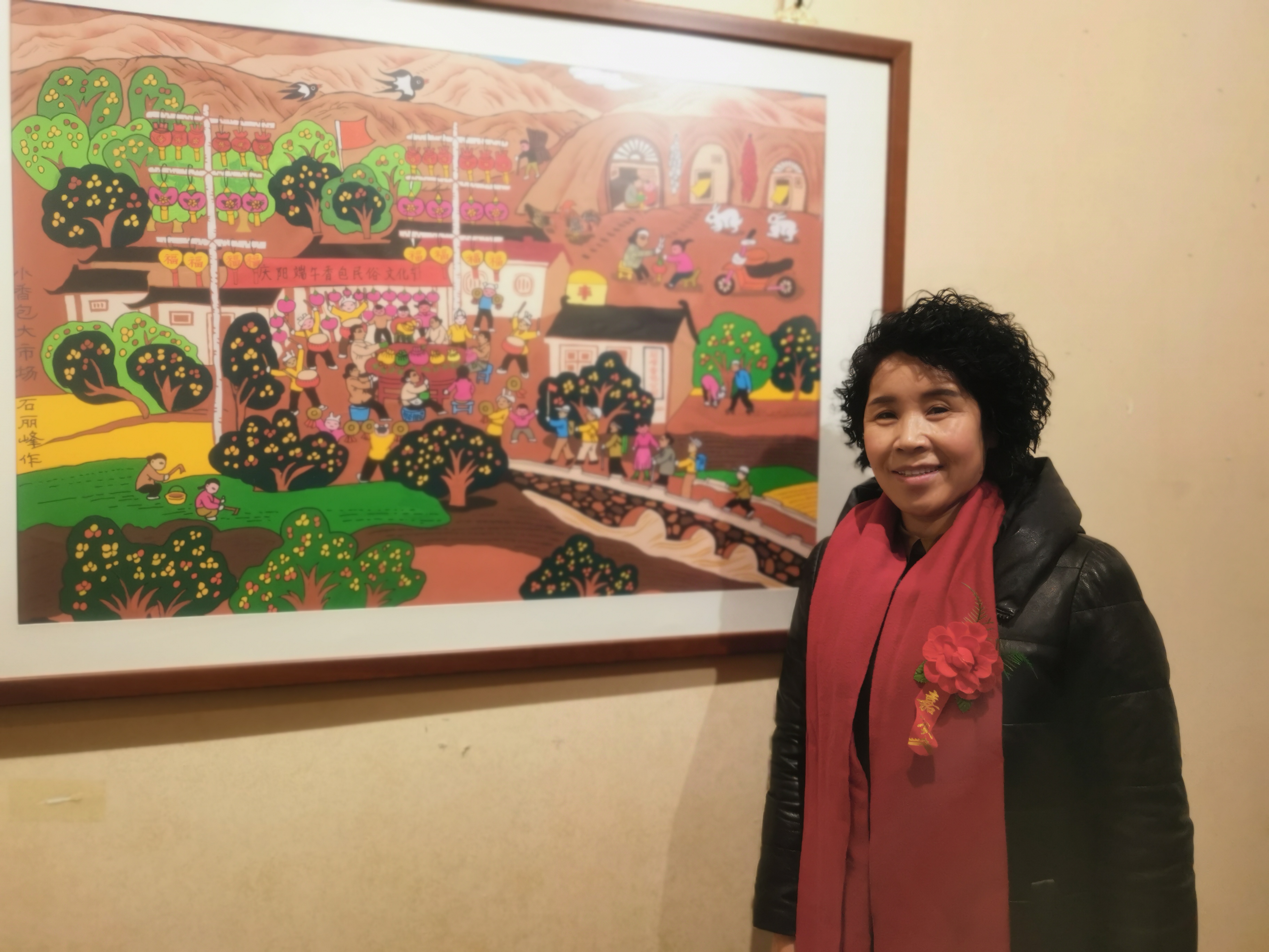 石麗峰的作品也參加了此次畫展，作為甘肅慶陽人的她既是一名農民畫家也是一名香包刺繡藝人。畫展上，石麗峰介紹，她的作品名為《脫貧致富的好產業》，最近才完成。反映的是一個農村婦女傳承香包製作工藝，全國各地的遊客前來觀摩，展現了黃土高原獨特的民俗文化和原生態的黃土風情。圖為石麗峰和她的作品《脫貧致富的好產業》。